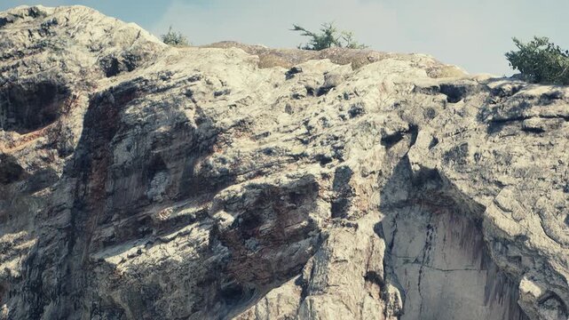 A large rocky cliff shows various textures and colors under bright sunlight. Sparse vegetation grows on top of the cliff. The setting reflects a natural landscape with clear skies.