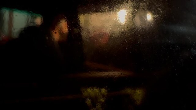 Blurred silhouettes of people moving through a city street at night, viewed through a car window, creating an atmospheric urban scene with motion, reflections, and low-light ambiance