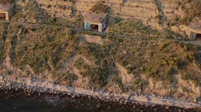Aerial view of a concrete bunker on a rugged cliffside with two people standing near the entrance in the Vjose-Narte Delta Protected Area in Zvernec, Vlore County, Albania.