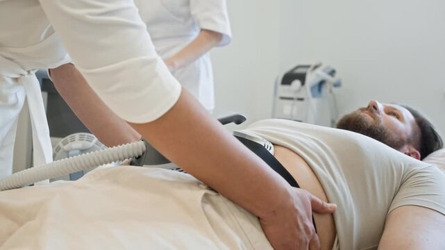 Caucasian man lying on clinic table while practitioners secure electromagnetic muscle stimulation device on his abdomen. Video perfect for medical spa and body contouring promos