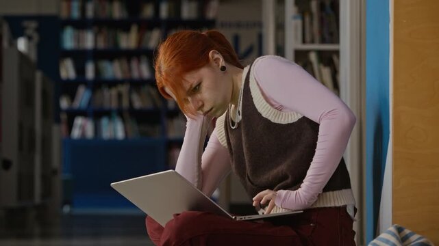Medium slow motion shot of Caucasian female teenager sitting on library floor, browsing laptop while studying. Clip for online education marketing, e-learning ads, and academic lifestyle campaigns
