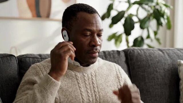 Middle aged african adult male person on sofa adjusting wireless earbud in cozy living room interior, relaxed listening focus and casual home comfort