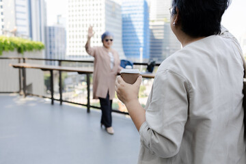 Fototapeta premium Senior Asian woman holds a coffee cup while looking at her friend on a city terrace, capturing urban recreation and relaxed friendship. reflecting friendship, active aging, and social connection