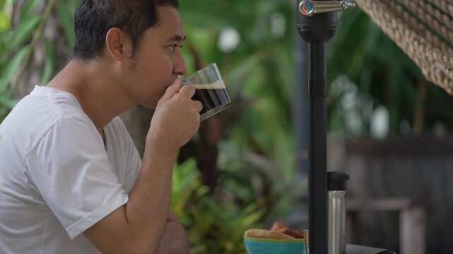Side profile of Asian man enjoying weekend morning routine, drinking coffee and checking news on smartphone while relaxing in peaceful home backyard garden. Static shot.