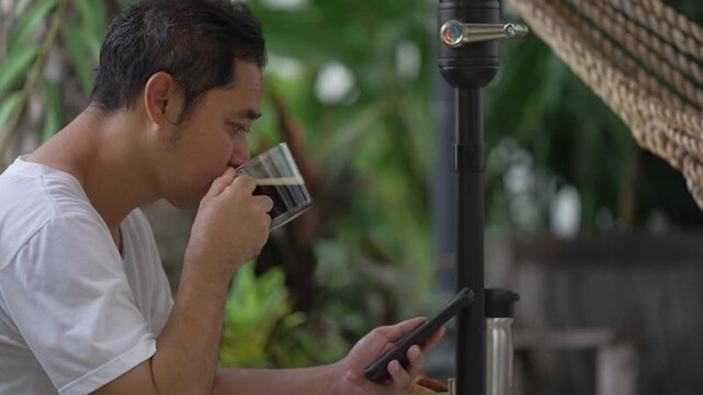 Focused Asian man deeply concentrating on smartphone screen while drinking coffee during his weekend morning routine in a peaceful backyard garden.