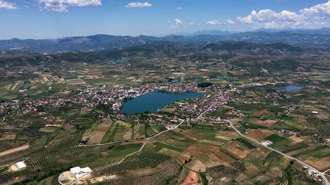 Aerial view of Lake Seferan surrounded by the town of Belsh and agricultural fields under a blue sky in Belsh, Elbasan County, Albania.