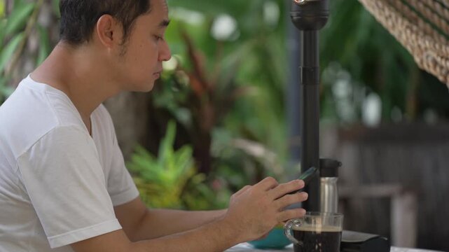 tilt up shot of Asian man enjoying weekend morning routine, drinking coffee and checking news on smartphone while relaxing in peaceful home backyard garden.