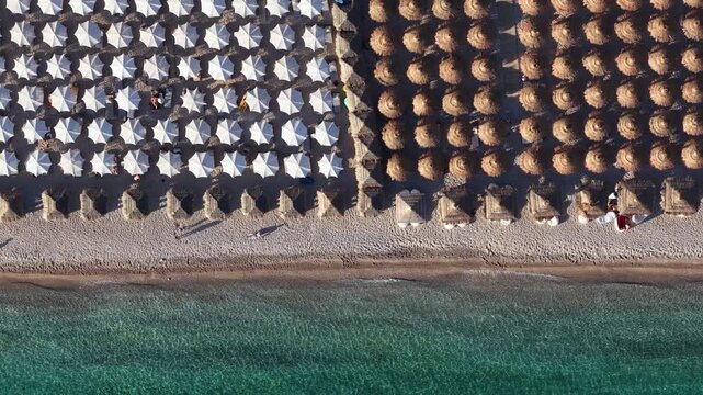 Aerial view of rows of white and straw umbrellas on the sandy shore of Llamani Beach with clear turquoise water in Himare, Vlore County, Albania.