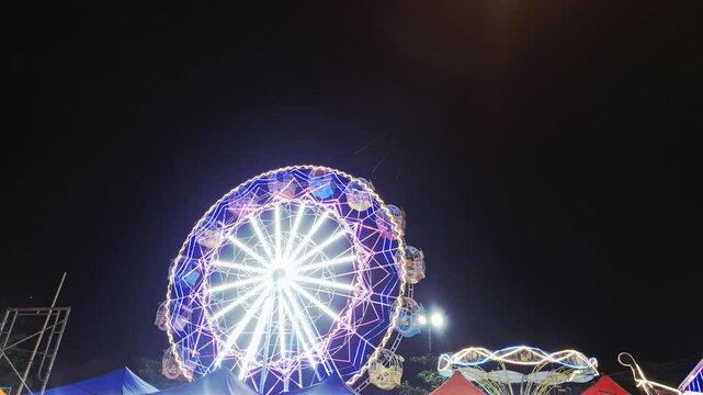 vibrant night scene at a carnival featuring a large, brightly illuminated Ferris wheel spinning slowly against the dark sky. The wheel is adorned with colorful LED lights, creating a captivating light
