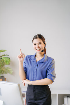Customer Service Professional: A friendly female customer service representative with a headset smiles and points upwards, offering assistance and guidance in a professional office setting.