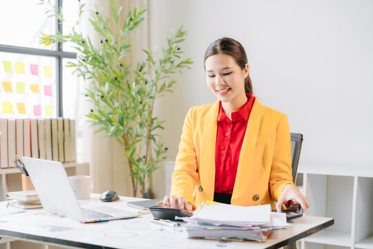 Focused Professional: A young Asian woman in a vibrant yellow blazer smiles while working diligently at her desk, surrounded by office supplies and a laptop.