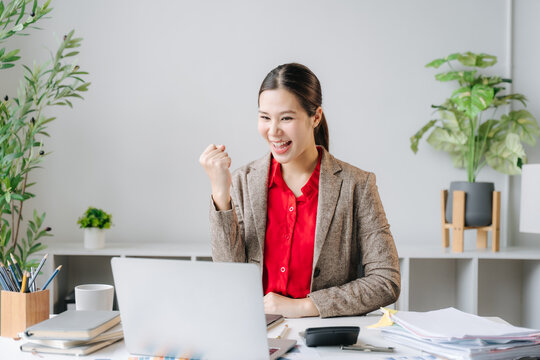 Woman Celebrates Success at Work: A young woman in professional attire smiles with joy and triumph, pumping her fist in the air as she looks at her laptop screen.