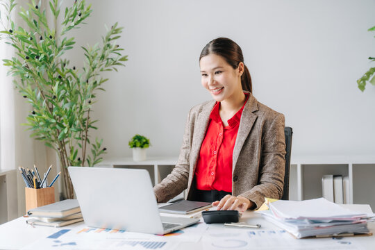 Focused Professional: A young woman in business attire smiles as she works diligently on her laptop at a well-organized desk, surrounded by office supplies and plants.