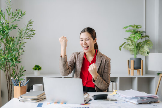 Excited businesswoman celebrating success at her desk. A young Asian woman, dressed in a smart blazer, cheers with her fists clenched as she looks at her laptop screen.