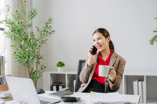 Businesswoman in Office: A young professional businesswoman smiles while talking on her mobile phone and holding a coffee cup in a modern office environment, with a laptop and plant in the foreground.