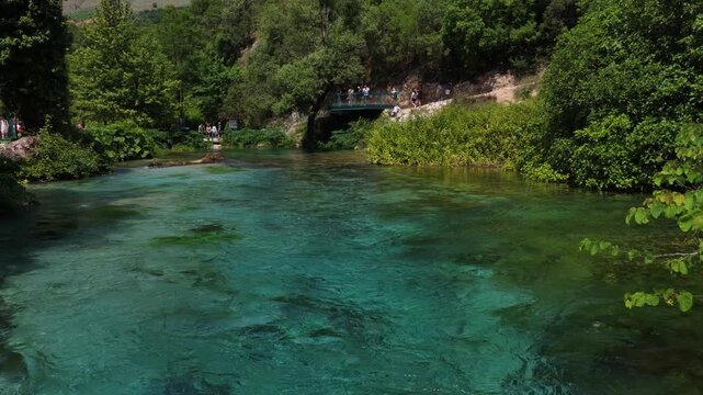 Aerial view of the Blue Eye spring with crystal clear turquoise water surrounded by lush green forest and tourists on a bridge in Finiq, Vlore County, Albania.