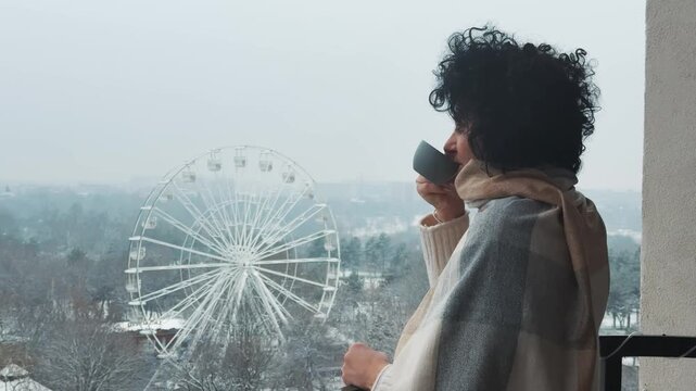 Woman Sips Hot Drink on Balcony Overlooking Snowy Bucharest Ferris Wheel
