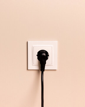 Black power plug connected to white electrical wall outlet on light beige wall. Simple and minimalist shot representing energy, connection, or utility