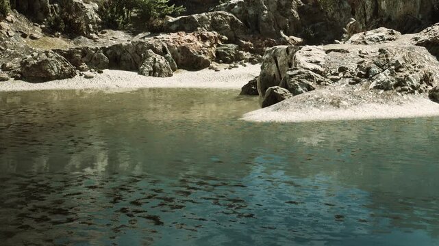 The scene shows a rocky shoreline with clear water reflecting the surrounding landscape. Pebbles and sand are visible along the edge. Greenery can be seen in the background.
