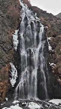 Cinematic vertical zoom shot of a beautiful waterfall cascading down rocky cliffs with frozen ice patches in winter.