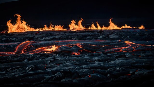 Molten lava stream flowing across dark volcanic landscape at night with flames in the distance.