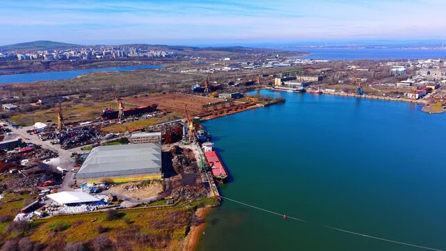 Industrial port area with scrap metal piles, Bulgaria. Wide drone view of cargo ship docked beside a warehouse and iron recycling yard under a blue sky.