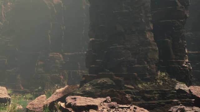 Tall rock formations rise from the ground in a canyon. Sunlight filters through the gaps, creating bright areas. Green grass grows among the stones. It is midday with clear light.