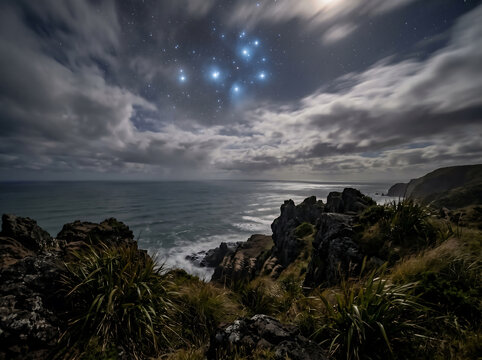 A dramatic night landscape of a rugged coastline with the ocean under a cloudy sky, illuminated by the moon and featuring the prominent Pleiades star cluster.