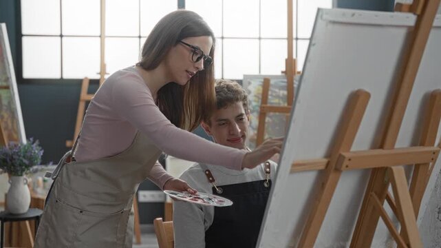 Woman painter in apron holds palette and guides man's hand with brush on canvas in studio, leaning over an easel to demonstrate; mentoring calm focus.