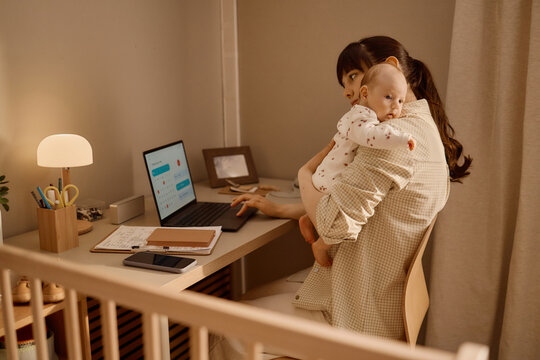 Caucasian mother holding baby while working on laptop at home, balancing childcare and remote job duties. Ideal for parenting, work life balance, maternity leave marketing