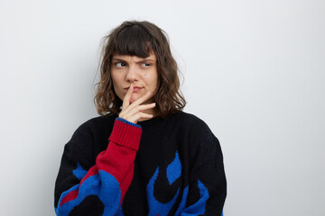 A thoughtful young woman stands against a light wall, finger resting on her lips as she contemplates a question. She wears a dark knit sweater with bright blue and red patterns, creating a moody, © SHOTPRIME STUDIO