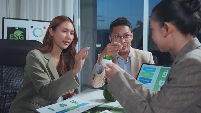Group of asian business people having an esg meeting to discuss corporate social responsibility and a green business strategy, reviewing data on a tablet and paper charts in an office at night