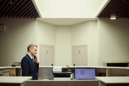 Wide shot of Caucasian middle aged hotel receptionist speaking on desk phone at front desk, managing guest service communication. Ideal for hospitality, travel booking, customer support