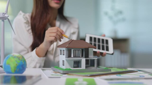 Female architect presenting a residential house model with solar panels to a client, discussing renewable energy solutions and sustainable living for a green and eco friendly home