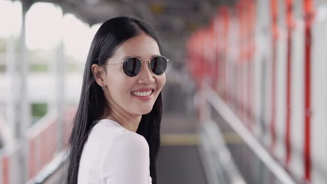 Over-the-shoulder shot of a cheerful young Thai woman wearing trendy sunglasses. She turns around, smiles brightly at the camera, and walks confidently on an urban skywalk at a public transit station.
