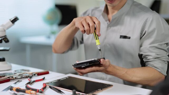 Professional technician using a screwdriver to unscrew a bolt on a computer hard disk drive while sitting at a desk with various tools and a disassembled tablet in a repair workshop