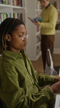 Vertical side view shot of African American teenage boy with glasses typing on computer in library while Caucasian teenage boy reading book in background. Video perfect for educational marketing