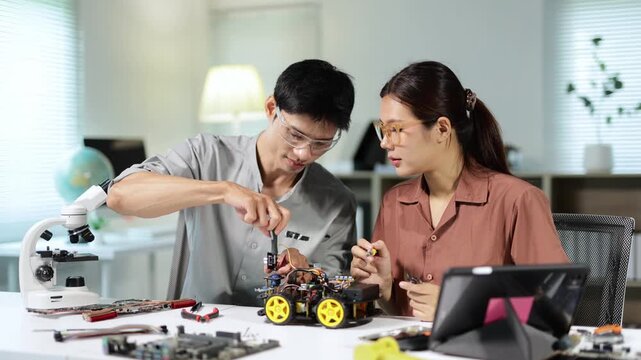 Two young Asian engineers in a modern laboratory are working together, assembling a small robotic car with screwdrivers and electronic components, discussing the innovative project with focus