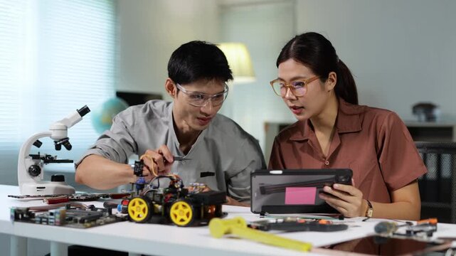 Young asian man and woman engineers cooperating on a robotics project, assembling and programming a remote controlled car model with a tablet in a modern university classroom or workshop