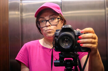 Woman photographer in pink cap and glasses with pouty expression taking mirror selfie with DSLR camera on tripod in elevator © Татьяна Швец