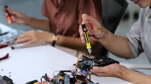 Male student technician using a screwdriver to assemble a custom robotic hard drive in a technology laboratory with another female student working on electronic components in the background