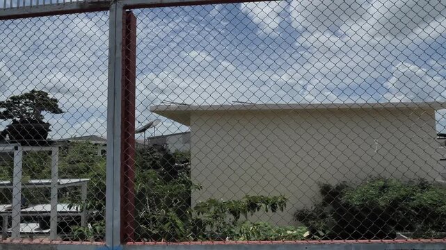 View of small beige building with roof and shrubs behind wire mesh fence under cloudy sky