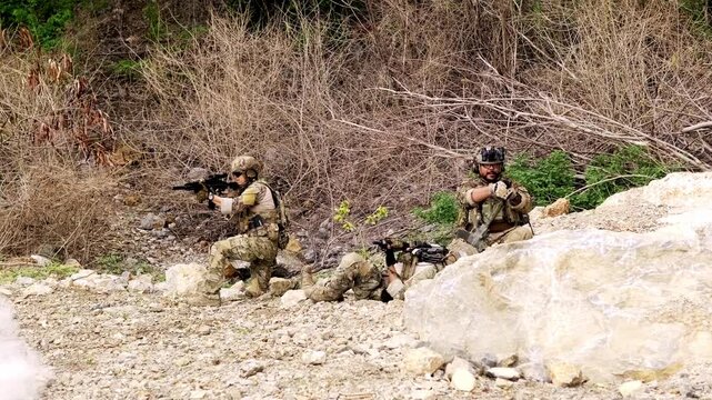 Three soldiers in camouflage navigate rocky terrain as two assist a fallen comrade during a tactical exercise in a dry, brushy landscape.