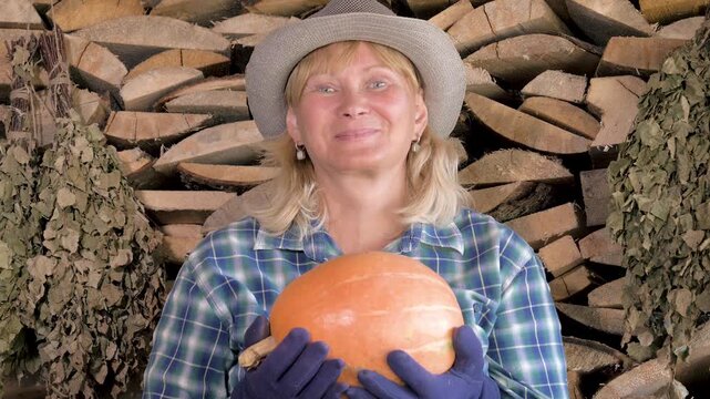 Happy blonde mature woman in plaid shirt and hat proudly holding large orange pumpkin from her garden harvest while standing in front of stack of firewood. Portrait female smiling at camera