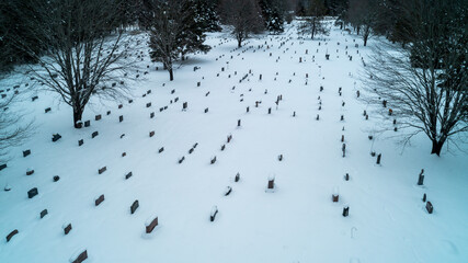 Fototapeta premium A high-angle drone photograph capturing an expansive cemetery covered in deep winter snow, with rows of granite headstones surrounded by dense pine trees on a cold, overcast day.