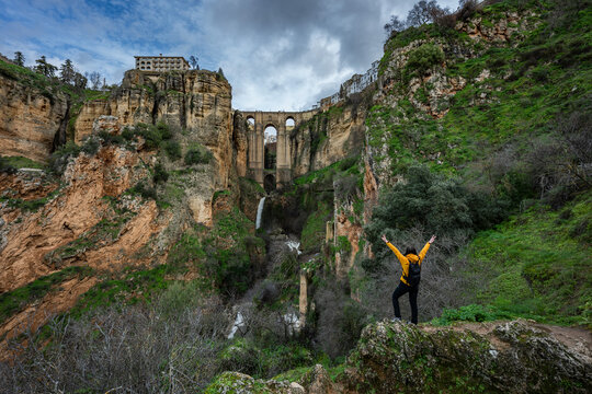 A traveler with a backpack standing on a rock with raised arms, overlooking the spectacular Puente Nuevo bridge and deep canyon in Ronda.