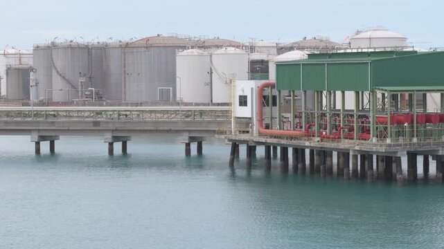 port fire protection station at 'map ta phut' port with pipe manifold, massive oil storage tanks, and a jetty over rippling turquoise water under a cloudy sky
