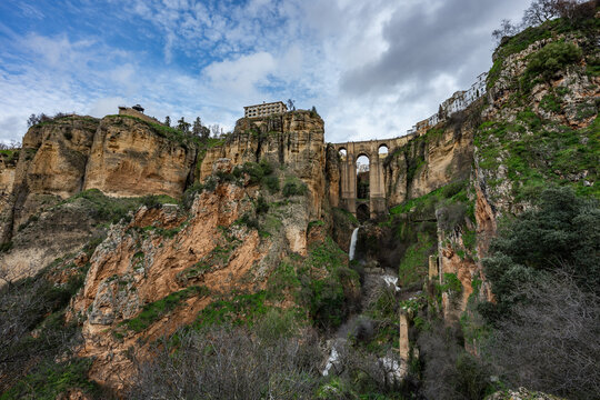 Wide angle view from the bottom of the gorge featuring the massive arches of Puente Nuevo bridge and a small waterfall in Ronda, Spain.