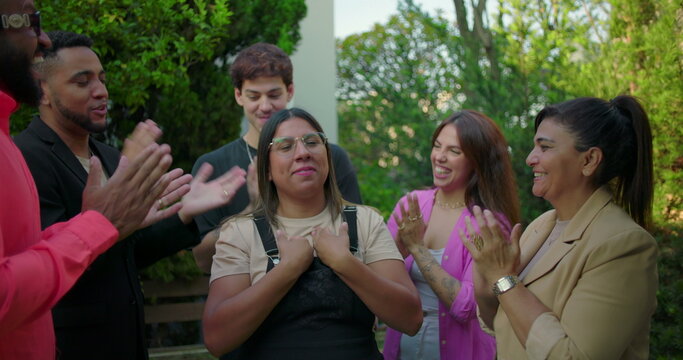 Latina woman celebrating at center of group as friends applaud her outdoors, expressing joy and excitement during heartfelt moment of recognition and support
