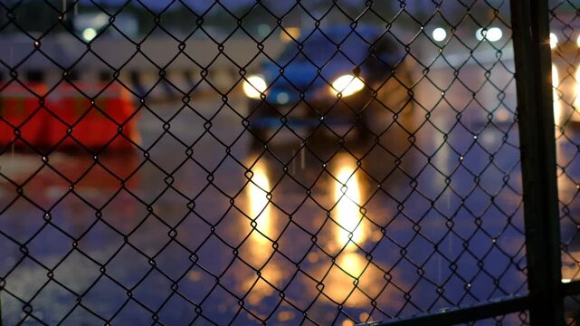 Night view through chain-link fence. Vehicles pass with glowing headlights. Rain-slicked road reflects lights. Urban scene with motion blur. Security barrier separates viewer from street